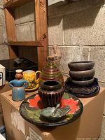 Overview of mixed ceramic decor including plates, bowls, vases, a jug, and garlic scraper arranged on a cardboard box in storage area