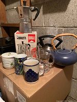 Bialetti Moka Express coffee maker with original box, six mugs, glass measuring cup, and two kettles on cardboard box.