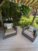Two chairs side by side on wood deck under striped awning showing grey wicker frames, beige cushions and striped pillows