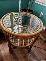 Front angled view of round rattan side table with circular glass top showing rattan frame and lower lattice-style shelf.