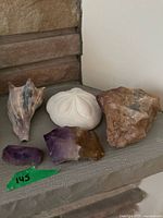 Five items displayed on a stone shelf: two raw amethyst pieces, one sand dollar, one conch shell, and one additional seashell.