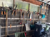 Wide view of seasonal garden and outdoor tools hanging on garage wall including rakes, shovels, hoes, pruning shears, and a wheelbarrow.