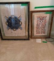 Both framed papyrus art pieces side by side, against a carpeted floor with visible labels on the frames showing measurements and origin.
