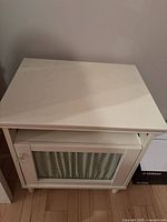 Front view of white bedside table showing hinged door with textured panel and crystal-like knob.