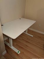 Photo showing white rectangular office desk with metal T-shaped legs and cable grommets in a corner of room with wood floor.