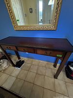 Dark brown wood console table positioned against blue wall beneath a large gold framed mirror.