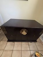 Front view of the dark brown wooden trunk showing the circular metal latch closure and metal corner reinforcements.