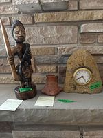 Front view of the wooden Yoruba warrior statue beside the small pottery vase and stone clock, all displayed on a shelf against a stone wall background.
