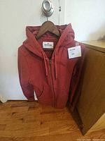 Front of maroon American Giant zip hoodie hanging, showing tag, zipper, pockets