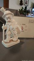 Front view of ivory glazed Little Cellist figurine beside original box