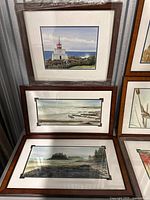 Three framed watercolor prints: lighthouse on shore, ocean with driftwood, lake and forest scene; displayed leaning against wall, plastic wrapped on one.