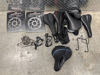 Top view of seven assorted bike saddles with varying shapes and padding, two brand new Supercycle disc brake rotors in original packaging, two black bottle cages, a used bike cassette, and a length of cable housing on a concrete floor.