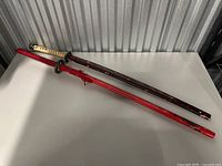 Two katana swords resting on table, showing full lengths with scabbards. Red and maroon colors with wear visible.
