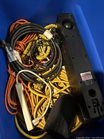 Bin containing a collection of power cables, extension cords, a black electronic device with a speaker grille or vent, a white power strip, yellow rope, and other miscellaneous accessories.