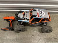 RC car side view showing body, tires, and orange roll cage next to remote controller on concrete ground.