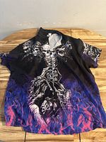 Black and purple short sleeve men's shirt with skulls and grim reaper on motorcycle graphic, laid flat on wooden table.