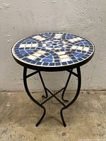 Small round table with a top in blue and white mosaic tile design and black wrought iron legs on a concrete floor in front of a textured wall.