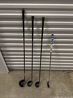 Four golf clubs standing upright against a wall: 3 metal woods and one putter with distinct grips.