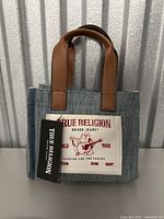 Front view of True Religion denim tote bag showing brown faux leather handles and branded patch with guitar player graphic.