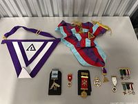 Photo of two sashes, one purple and white triangular with Masonic symbol, and one multicolor silk with attached pendants, alongside a collection of pins and medals arranged below them