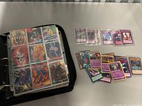 Black zip binder open to a 3×3 page showing Marvel cards (Ghost Rider, Thor, Spider-Man, Captain America, Blade) and loose Yu-Gi-Oh/Pokemon cards on table.