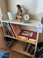 Full view of the bookshelf loaded with assorted books, ceramic planter, milk glass vase, vintage clock, and kitchen scale.