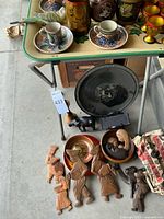 Lot overview showing cups, saucers, bird figurine, carved wooden figures, black platter, and wooden tray.