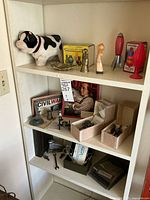Overview of lower three shelves of a bookcase showing various items including ceramic cow bank, figurines, historic miniature artillery cannons in boxes, vintage Rolodex, stapler, scissors, and other small metal items.