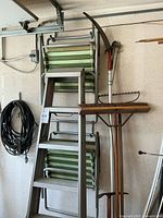 Four stacked green and white striped metal lawn chairs next to a 5-step aluminum folding ladder hung on the wall with a wood handle broom, metal rake, and curved scythe blade.