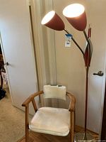 Photo showing the lamp with two cone-shaped red shades and brass accents next to the wooden armchair with white vinyl upholstery.