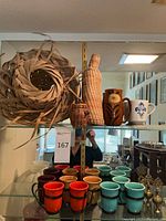 Colorful ceramic mugs in red, yellow, blue, brown with metal handles, woven decor including bottle cover and basket, arranged on glass shelf with mirror background.
