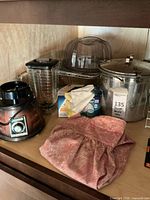 Two vintage Oster blenders, a plastic portable cake stand with lid, large metal pressure cooker, pink cloth towel, and boxed light bulbs all displayed on a wood shelf.