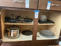 Open kitchen cabinet shelf showing stacked nonstick pans, Farberware pot, glass serving trays and baking trays.