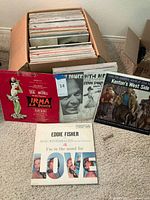 Photo of multiple vinyl LP album covers standing in front of a box containing more records. Visible albums include 'Irma La Douce,' 'Come Dance with Me' by Frank Sinatra, 'Kenton's West Side Story,' and 'I'm in the Mood for Love' by Eddie Fisher.