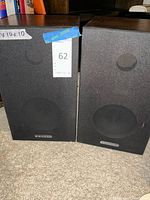 Pair of black vintage bookshelf speakers with fabric grills and visible speaker outlines, sitting on carpet.
