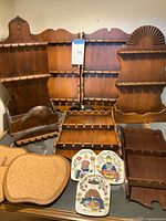 Six wooden spoon racks of varying sizes and styles displayed vertically and horizontally, along with four ceramic trivets on a table, including apple-shaped cork trivets.