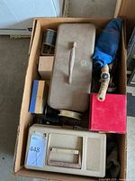 Top-down view of cardboard box containing two vintage fishing tackle boxes, spool of fishing line, fishing rod handle visible, and multiple small boxes likely with fishing accessories.