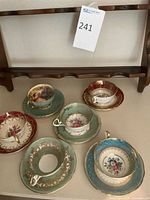 Wooden wall rack with six cup holders shown above six Aynsley fine china cups and saucers arranged underneath on a surface.
