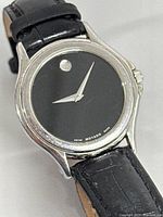 Close-up of Movado black dial wristwatch showing signature single dot at 12 o'clock, silver hands, stainless steel case, and black leather strap.