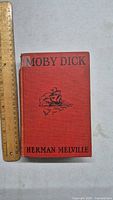 Front cover of 1925 Moby Dick book showing red cloth cover with title, author, and ship illustration in black.
