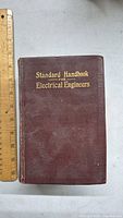 Front cover of the 1933 Standard Handbook for Electrical Engineers featuring gold embossed title on brown cloth cover with measuring ruler for scale.