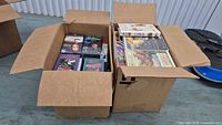 Two open cardboard boxes filled with assorted VHS tapes, viewed from above showing various tape cover art.
