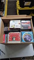 Top-down view of an open cardboard box filled with assorted music CDs of various artists and genres.