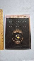 Front cover of 'The Universe In a Nutshell' hardcover book by Stephen Hawking with a ruler for size reference.