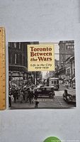 Front cover of 'Toronto Between the Wars' photography book showing a busy Toronto street scene from 1919-1939 with vintage cars and pedestrians.