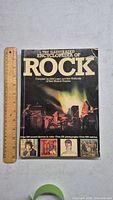 Front cover of The Illustrated Encyclopedia of Rock book showing title, author, and a concert photo with ruler for size reference.