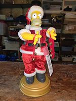 Front view of Homer Simpson figurine dressed as Santa Claus, standing on gold base, showing red velvet suit with white trim and jingle bells.