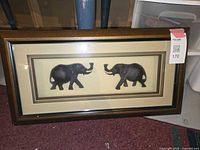 Front view of framed shadow box art featuring two black elephant 3D figures facing each other, set against beige background with multi-bordered frame in dark brown and gold.