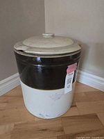 6 gallon two tone crock with lid sitting on wooden floor, showing full crock and lid with visible chips on lid edge.