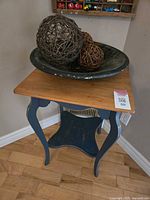 Wood accent table with natural wood top and dark blue curved legs and lower shelf, shown with decorative items on top.